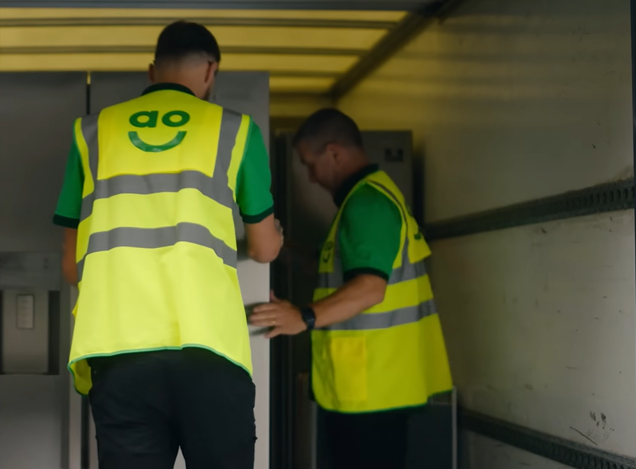 Two AO delivery drivers wearing high-visibility vests load a customer’s old fridge into an AO delivery vehicle for recycling.